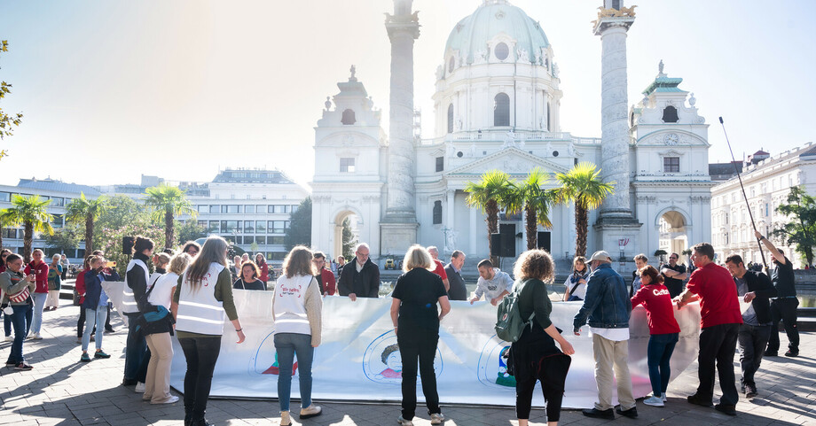 Eine Menschengruppe hält ein großes Transparent in den Händen und steht vor der Karlskirche