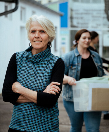 Eine Frau mittleren Alters steht mit verschränkten Armen. Im Hintergrund tragen Personen Kisten.